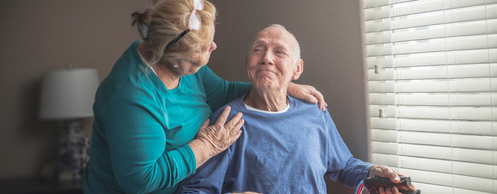 n elderly man sitting by a window, holding a cane, and smiling warmly at an elderly woman who is standing beside him, gently resting her hand on his shoulder.