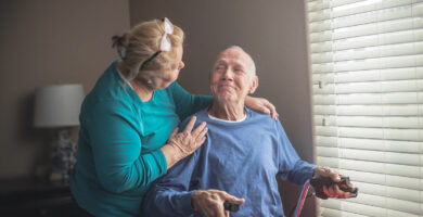 n elderly man sitting by a window, holding a cane, and smiling warmly at an elderly woman who is standing beside him, gently resting her hand on his shoulder.