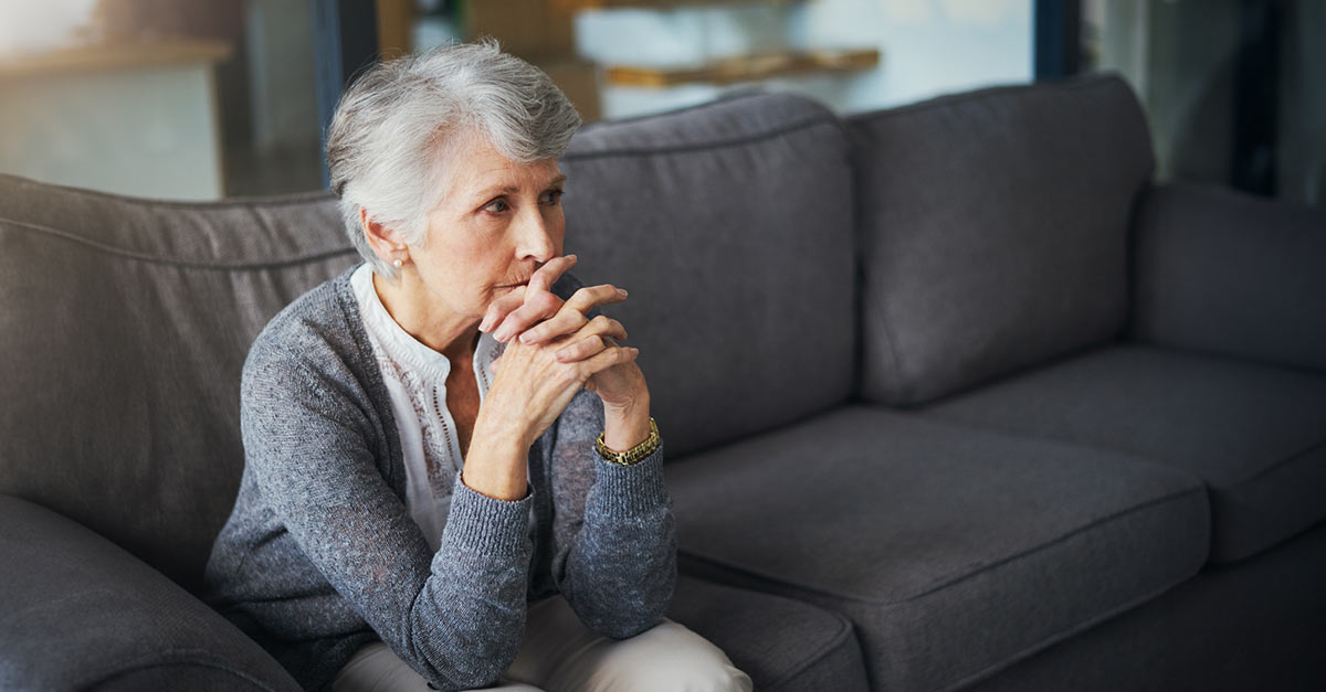 Elderly woman sitting on a couch, deep in thought, with her hands clasped in front of her face