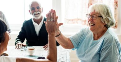 An older woman high fives another older woman sitting at a kitchen table