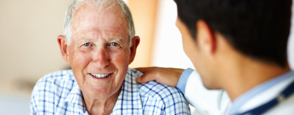 A doctor reassures an older man who's smiling into the camera.